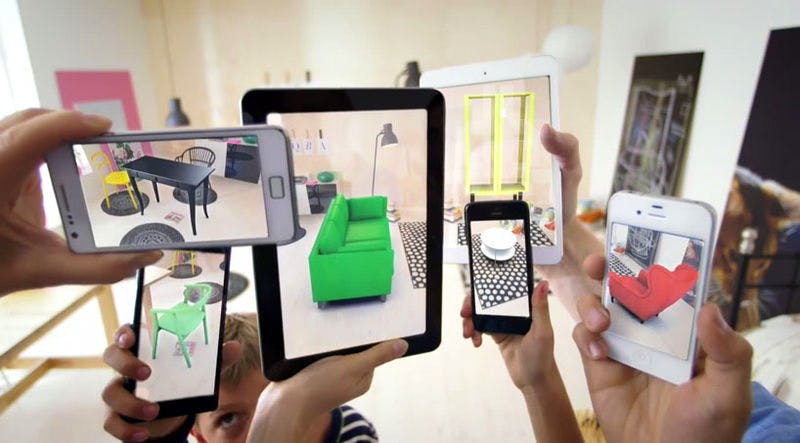 Several hands hold up smartphones, which are projecting different items of furniture into a room via augmented reality.